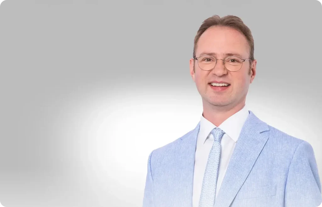 Rechtsanwalt Robin Schmid im hellblauen Anzug vor neutralem Hintergrund, freundliches Portrait